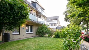 A residential building with a well-maintained lawn, a balcony with glass railings, and a small garden area with various plants. - Wellenklang – Your Cozy Vacation Rental Near the Sea (Büsum)