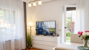 A modern living room with a television, a dining table, a white sofa, and a view of the outdoors. - Wellenklang – Your Cozy Vacation Rental Near the Sea (Büsum)