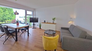 En moderne stue med en grå sofa, en gul lænestol, et spisebord med stole, et fladskærms-tv og en lille potteplante.