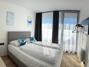 Une chambre d’hôtel moderne avec un lit aux lignes courbes, du linge de lit blanc et une grande fenêtre agrémentée de rideaux légers.