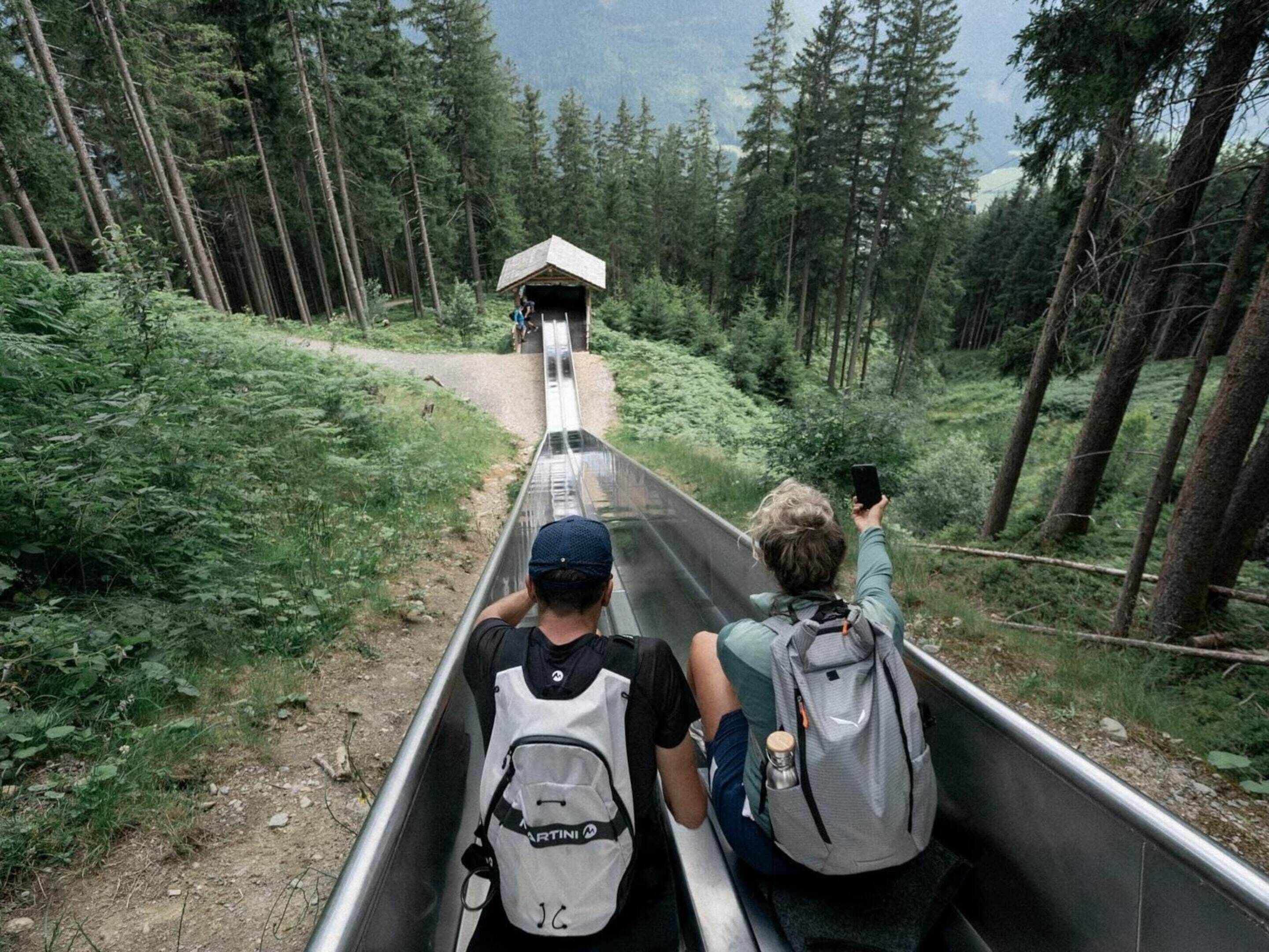 A person in a metal slide with a backpack, surrounded by a forested area.