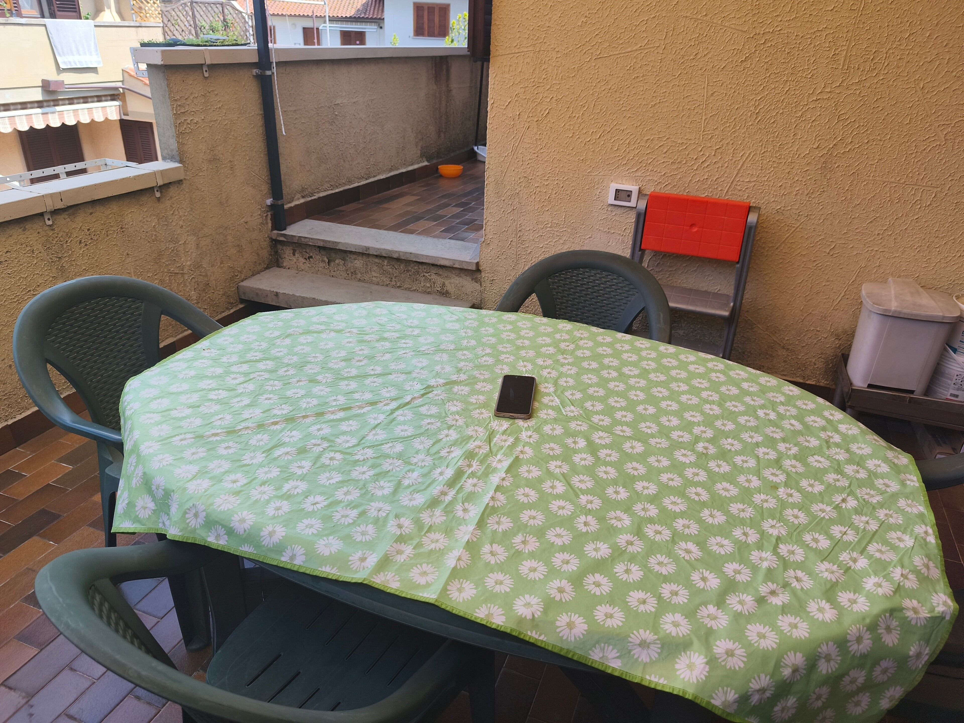 Eine kleine Außenveranda mit einem grünen Tisch und zwei Stühlen, einer roten Mülltonne und einer weißen Steckdose an der Wand.