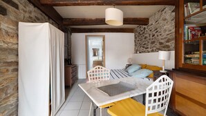 A room with a stone wall, a bed with yellow pillows, a white table with a mat, a white chair, a wooden cabinet with books, and a hanging light fixture.