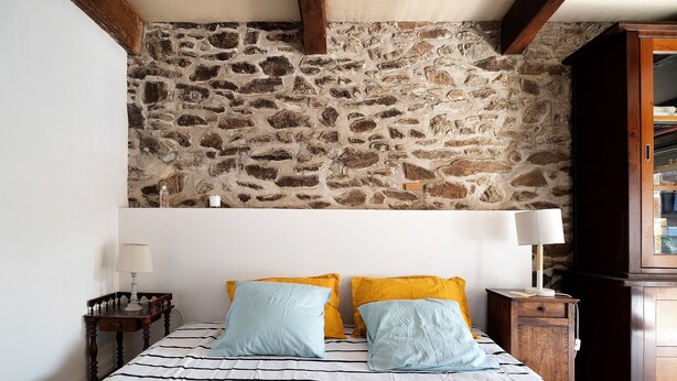A bedroom with a stone wall, a bed with striped bedding, two pillows, a nightstand with a lamp, and a wooden cabinet.