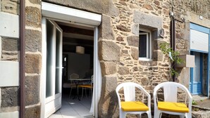 A stone building with a door leading to an interior space, two white plastic chairs with yellow cushions outside, and a small window with a plant.