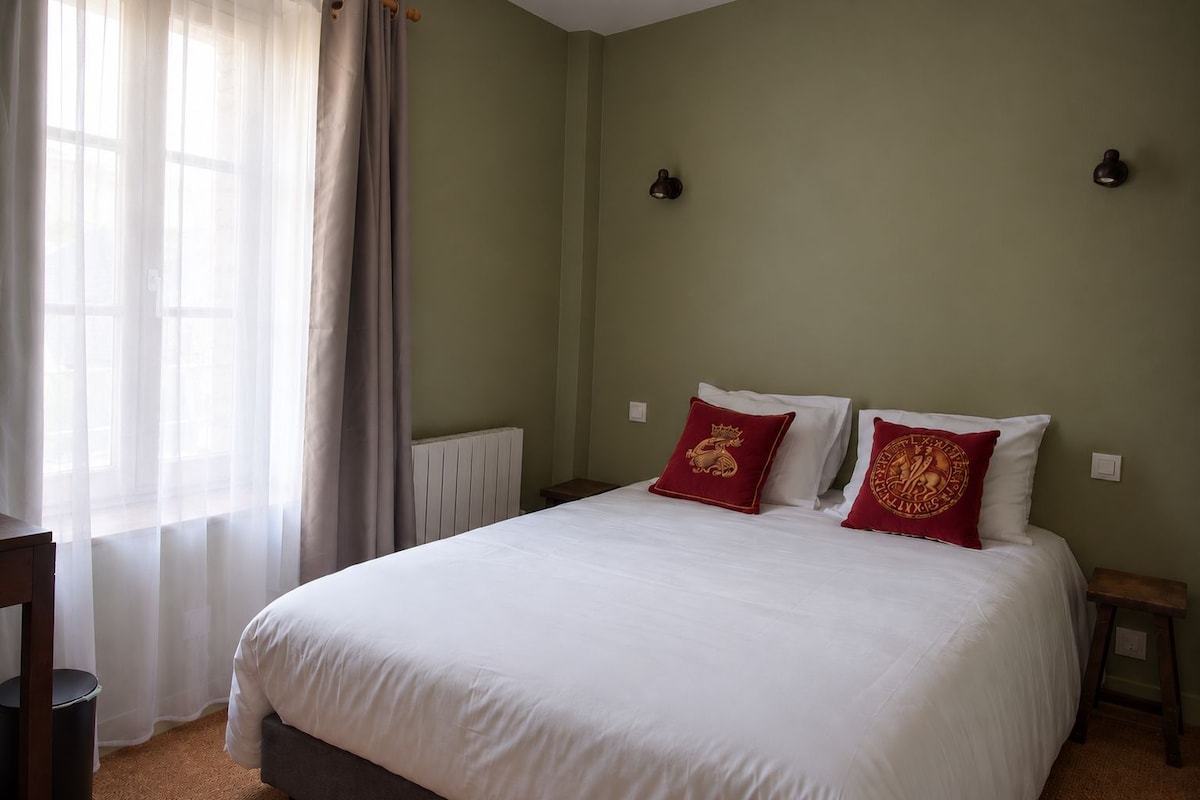 A bedroom with a large bed, two pillows with a crest design, a radiator, a bedside table, and a window with sheer curtains.