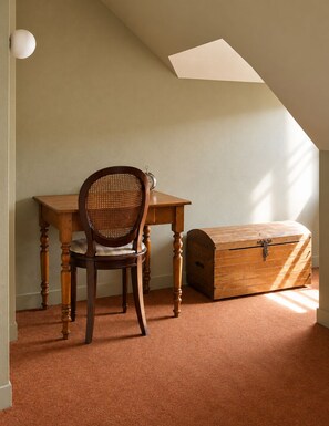 A room with a wooden desk, a wicker chair, a wooden chest, and a sloped ceiling with a skylight.