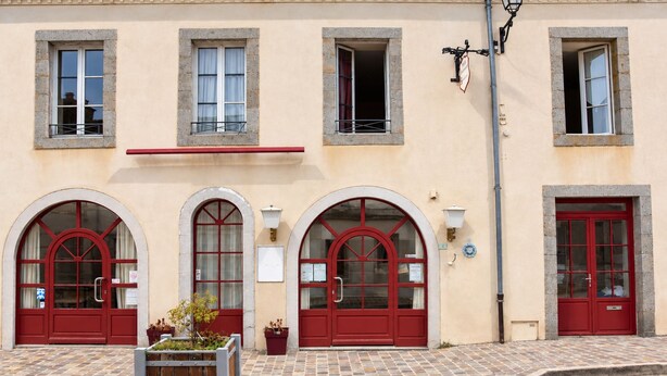 A building with three arched windows and red doors, a tiled pavement, and a small planter with plants.