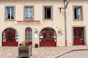 A building with three arched windows and red doors, a tiled pavement, and a small planter with plants.