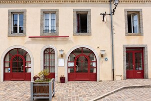 Un edificio con tres ventanas arqueadas y puertas rojas, pavimento de baldosas y una jardinera con plantas.