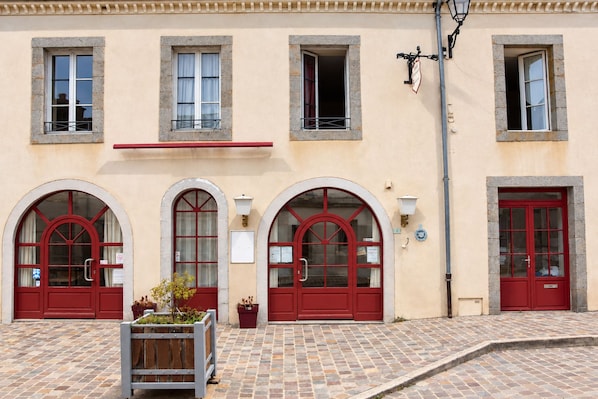 A building with three arched windows and red doors, a tiled pavement, and a small planter with plants.