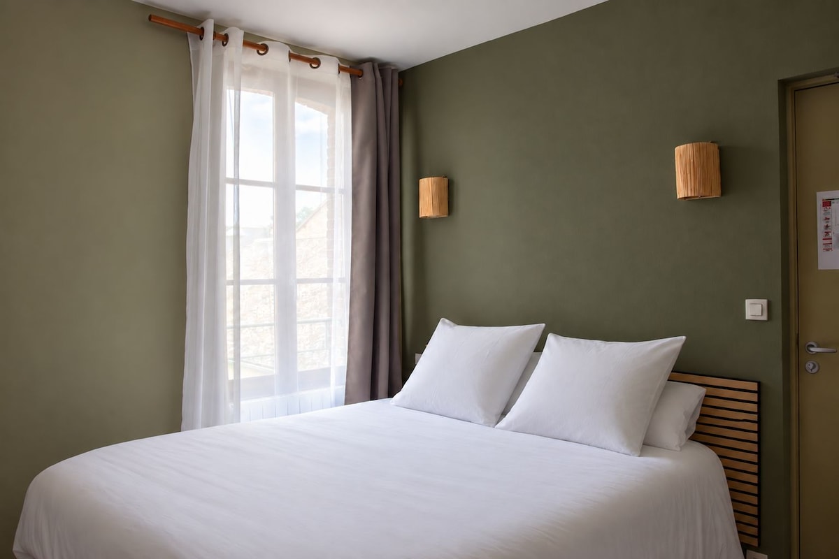 A hotel room with a large bed, a window with curtains, and a wall-mounted lamp.