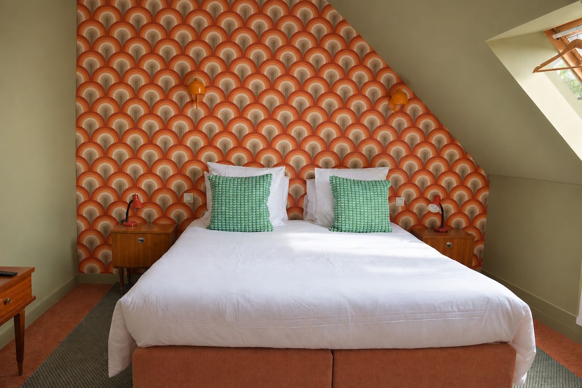 A bedroom with a bed, two bedside tables, a patterned wallpaper, and a sloped ceiling.