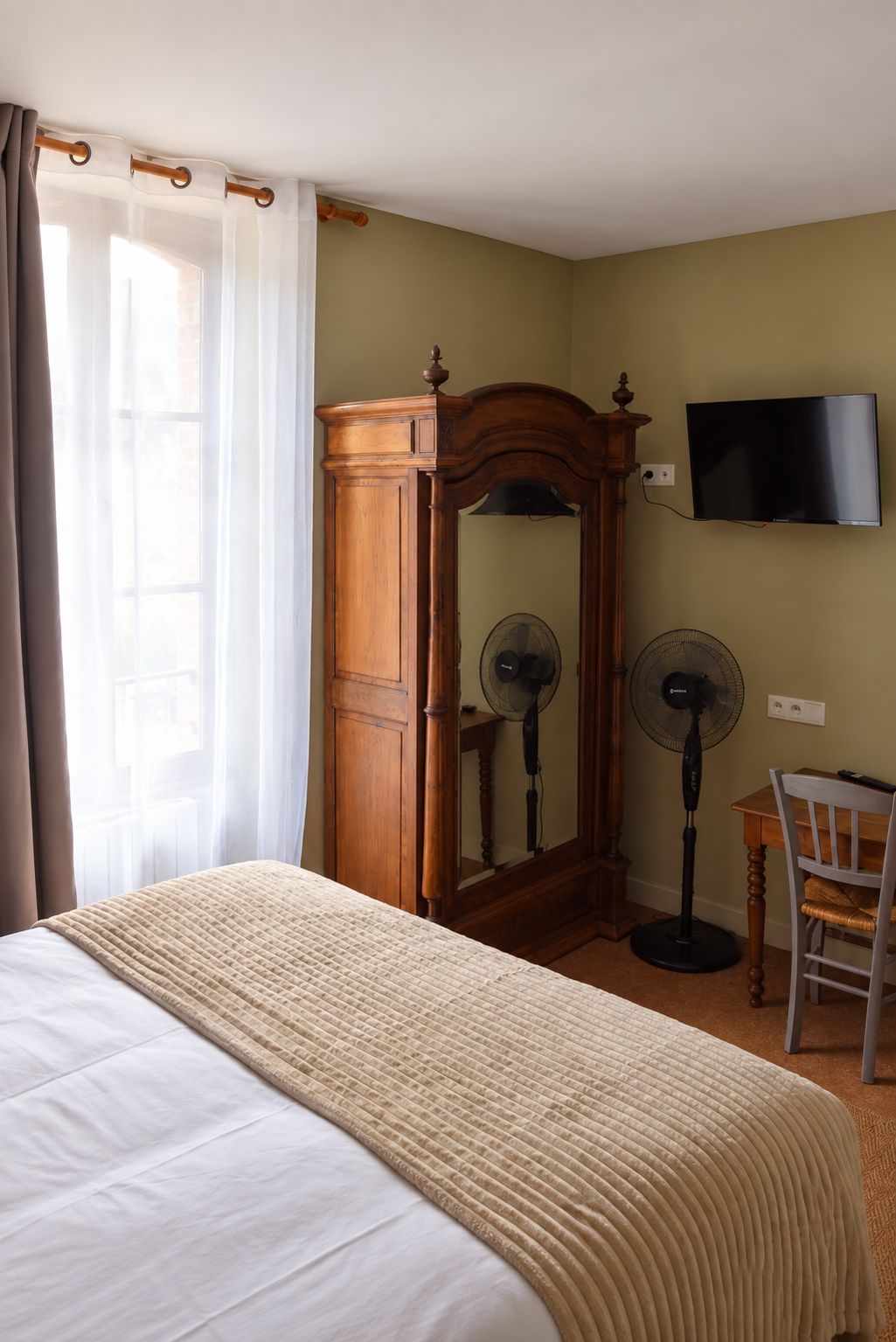 Un dormitorio con una cama, un armario de madera, un espejo, dos ventiladores, un televisor y una silla.