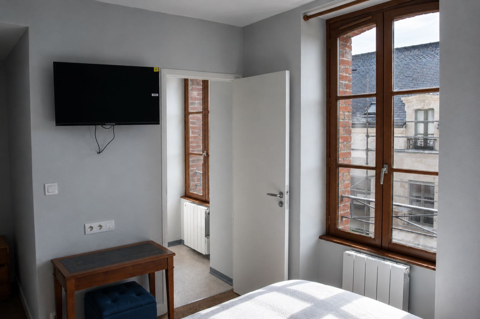 Une chambre à coucher avec un lit, un téléviseur fixé au mur, une fenêtre donnant sur des immeubles et une porte menant à une autre pièce.