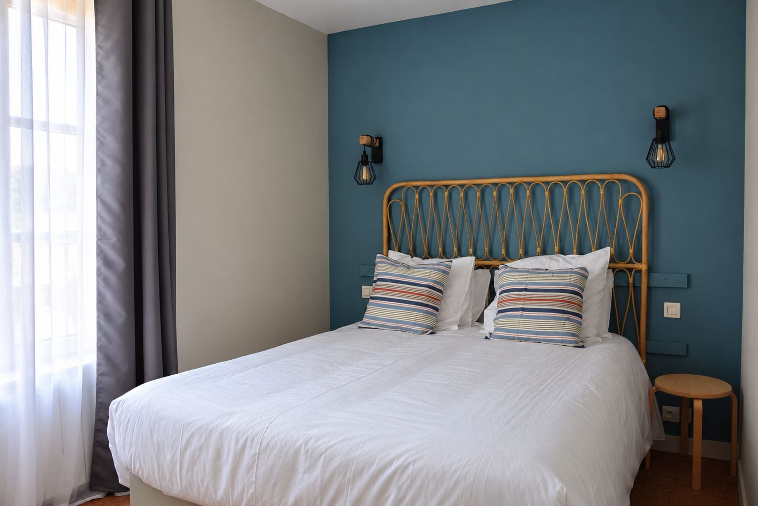 Una cama con colchón blanco y almohadas a rayas, cabecera de madera y dos lámparas fijadas a la pared.