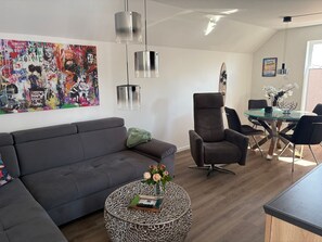 A modern living room with a grey sofa, a round coffee table with a book and flowers, a grey armchair, and a glass dining table with chairs. - Klabauter-Kajüte - Balcony, private parking space, washing machine, infrared cab (Olpenitz)