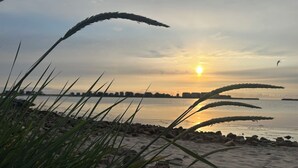 Una playa con rocas y hierba en primer plano, un atardecer al fondo y un horizonte urbano distante.