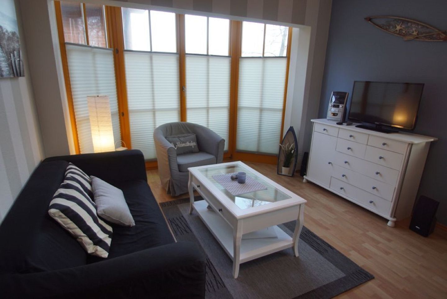 Una sala de estar con un sofá negro, un sillón gris, una mesa de centro de vidrio, un aparador blanco y un televisor.