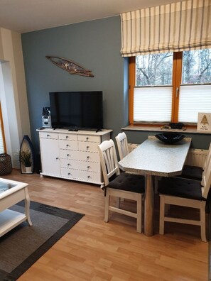 Ein Zimmer mit Esstisch, Stühlen, einem Fernseher auf einem Schrank, einem Fenster mit Jalousien und einer dekorativen Seeanemone.