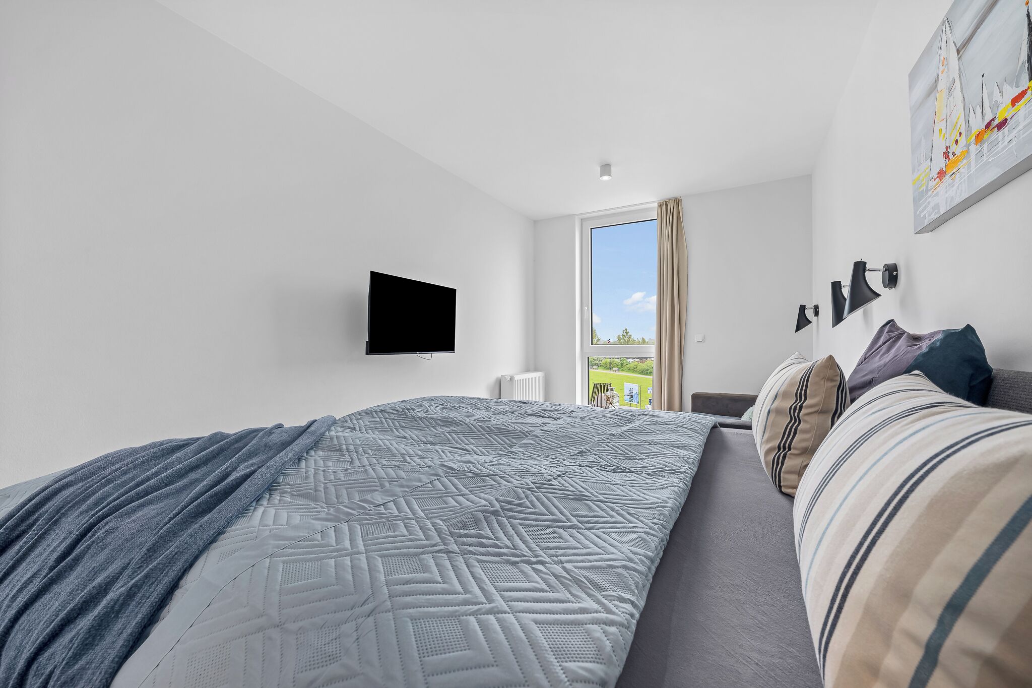 A modern bedroom with a bed, a flat-screen TV, a window with a view, and a painting on the wall.