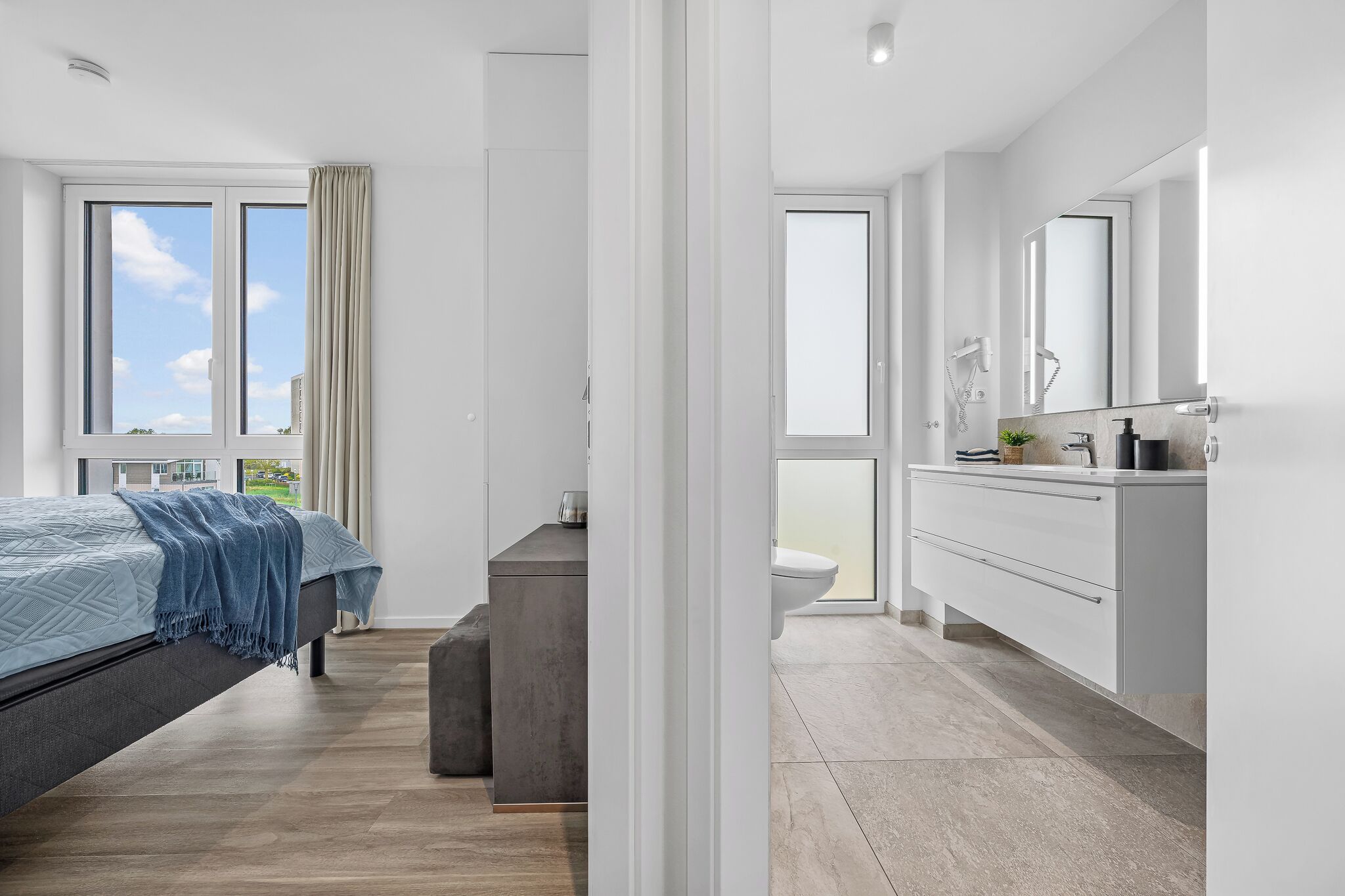 Une chambre à coucher moderne avec un lit, une table de chevet et une vue sur l’extérieur. L’image montre également une salle de bain avec des toilettes, un lavabo et un miroir.