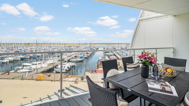 A balcony with a view of a marina, wicker furniture, a vase with flowers, and a menu. - Spacious Apartment with exceptional Sea View and Wellness Area! (Wendtorf)