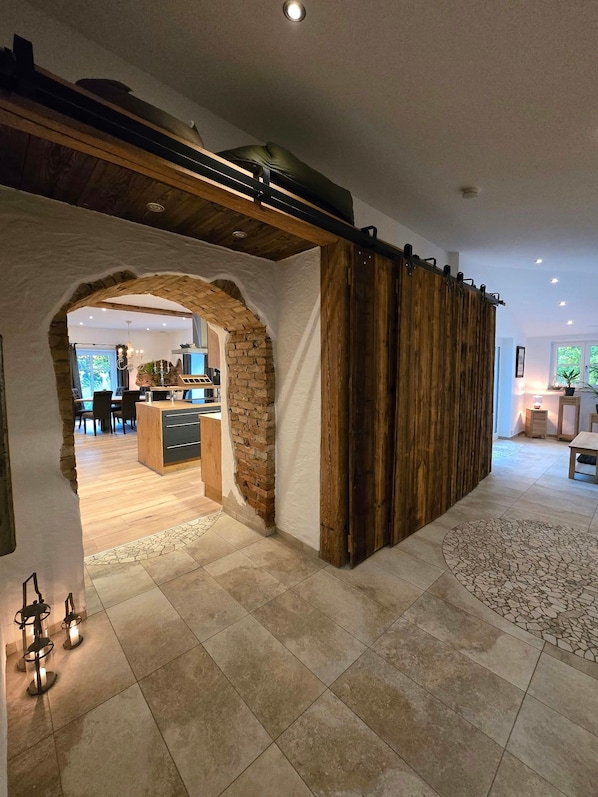 A modern interior with a stone archway leading to a kitchen and dining area, a wooden sliding door, and a tiled floor.