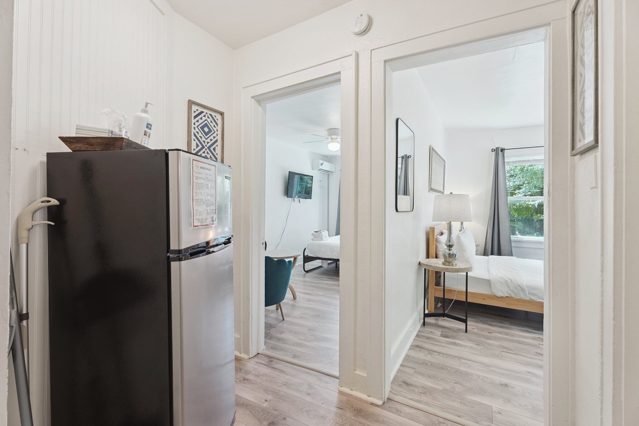 A modern hotel room with a wooden floor, a bed with a white comforter, a bedside table with a lamp, and a view of greenery outside.