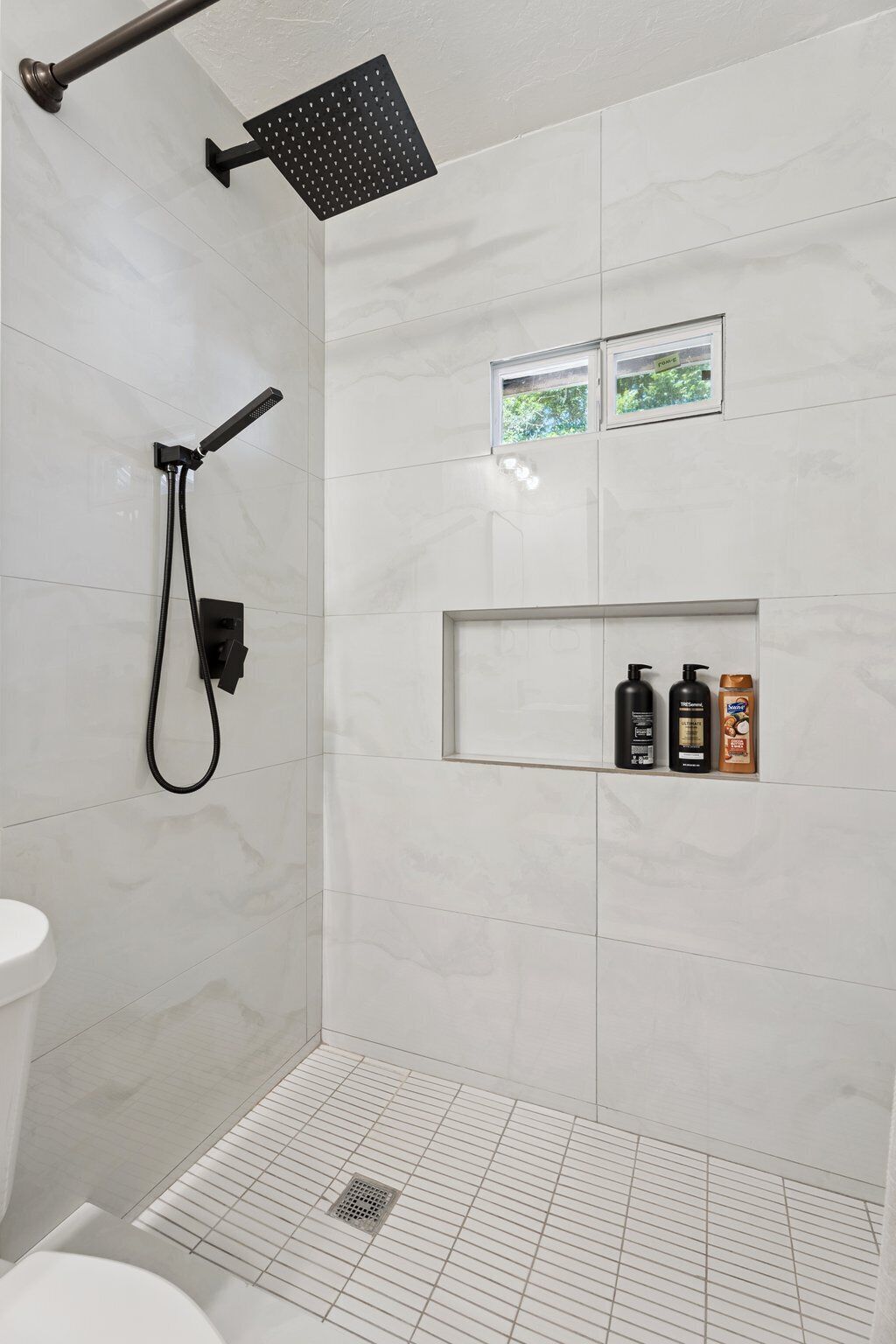 Un espace douche moderne doté d’un pommeau de douche à main, d’un panneau de commande mural et d’une petite étagère pour les articles de toilette.