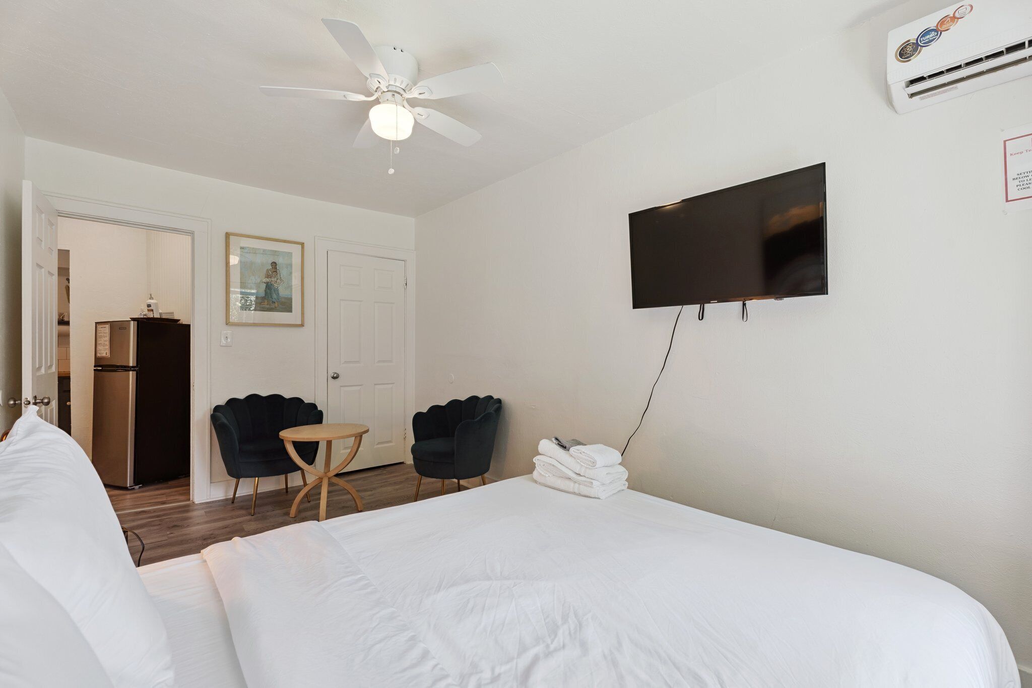 Een hotelkamer met een bed, een plafondventilator, een televisie, een kleine tafel, twee stoelen en een koelkast.