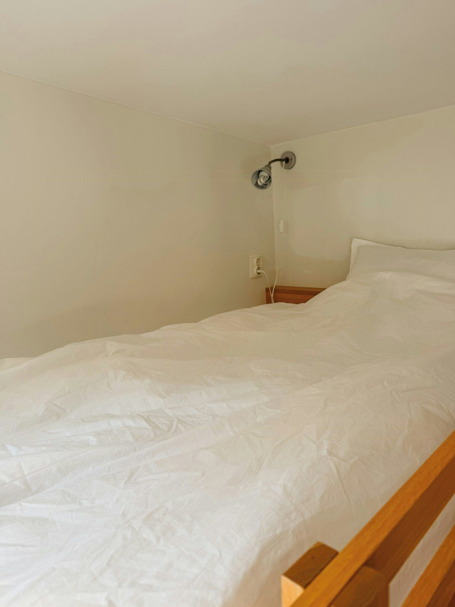 Uma cama de solteiro com cabeceira de madeira em quarto com paredes lisas e luminária no teto.