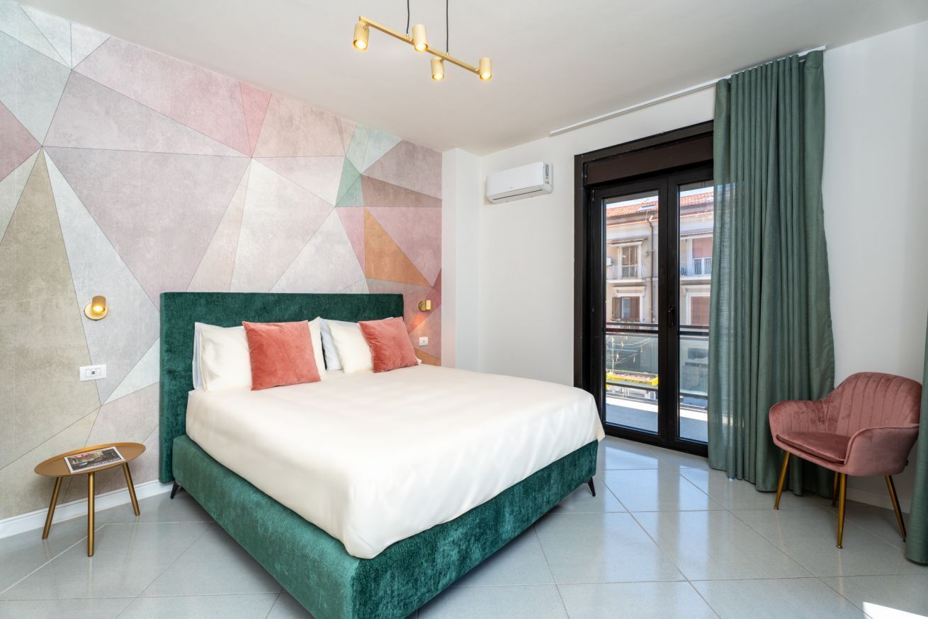 A modern hotel room with a large bed, a green headboard, a small side table, a pink chair, and a geometric-patterned wall.