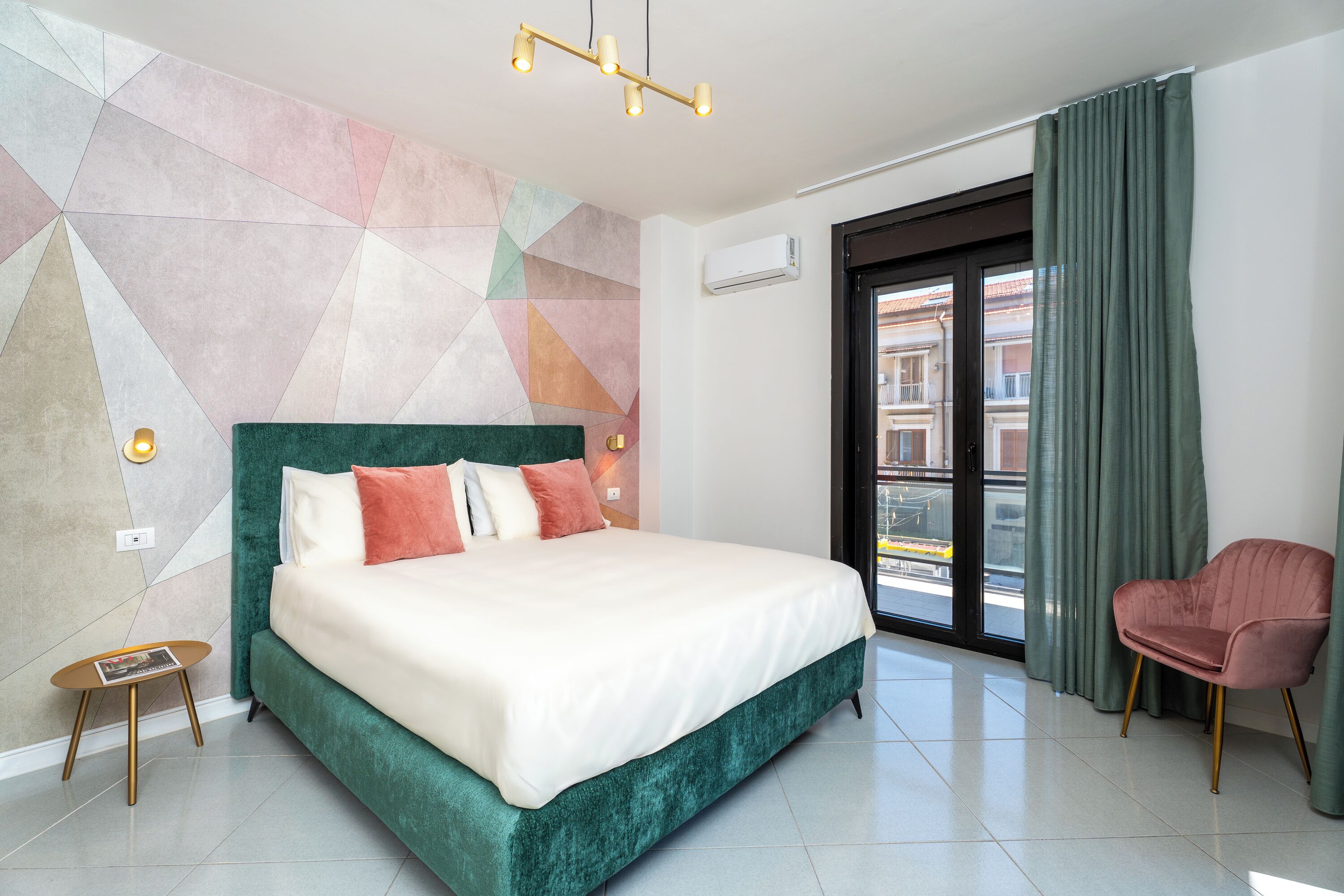 Une chambre d’hôtel moderne avec un grand lit, une tête de lit verte recouverte de tissu, une petite table d’appoint, une chaise rose et un mur orné d’un motif géométrique.