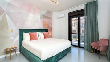 Une chambre d’hôtel moderne avec un grand lit, une tête de lit verte recouverte de tissu, une petite table d’appoint, une chaise rose et un mur orné d’un motif géométrique.