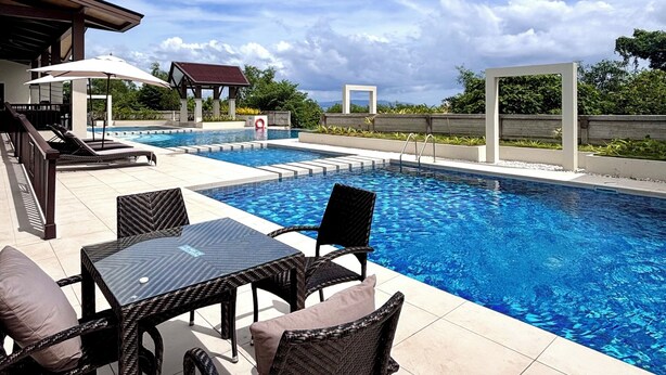 Ein Schwimmbecken mit Liegestühlen und Sonnenschirmen, umgeben von einer gefliesten Terrasse und viel Grün.