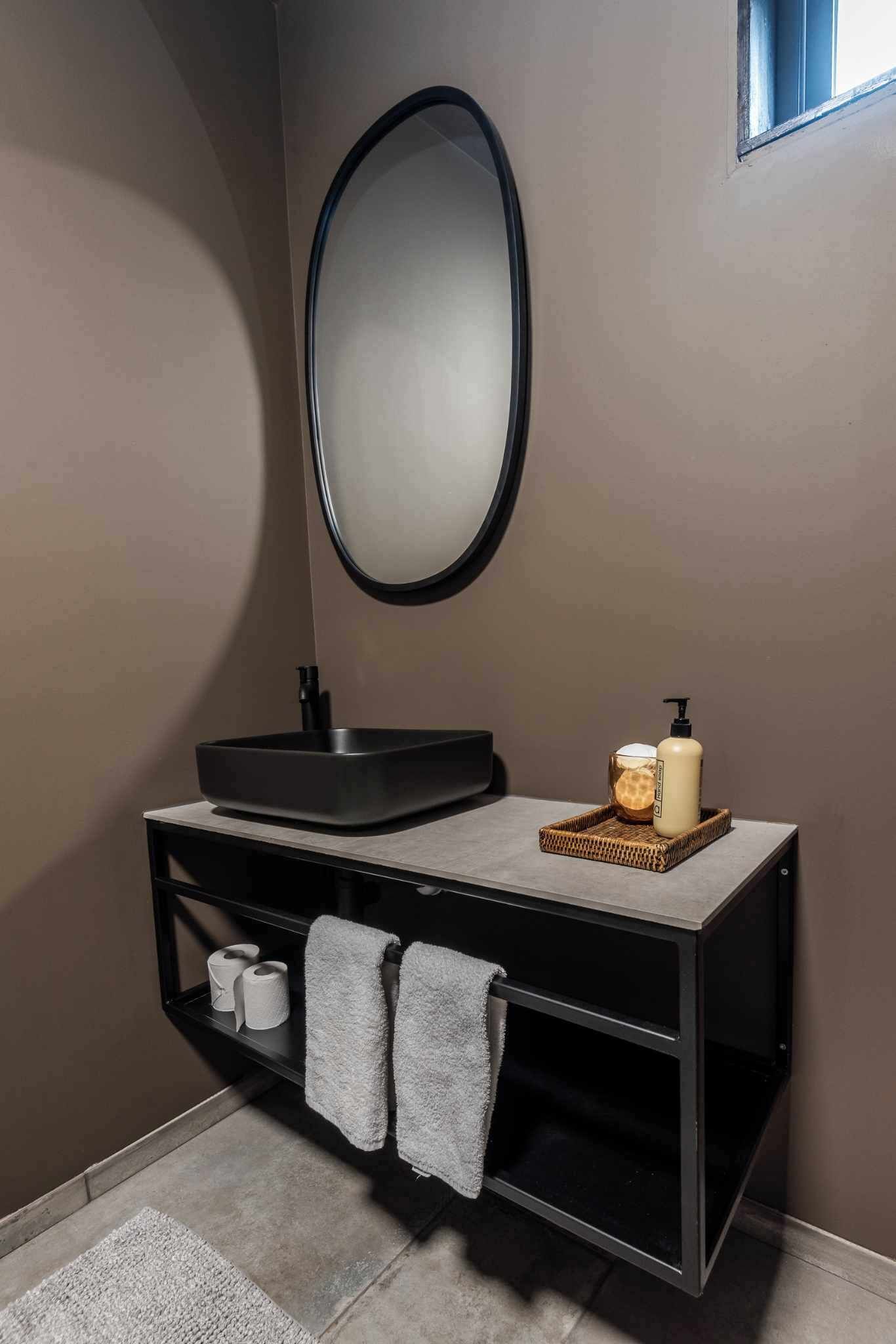 A bathroom with a black vanity, a round mirror, a black sink, two towels, a soap dispenser, and a bottle of lotion.