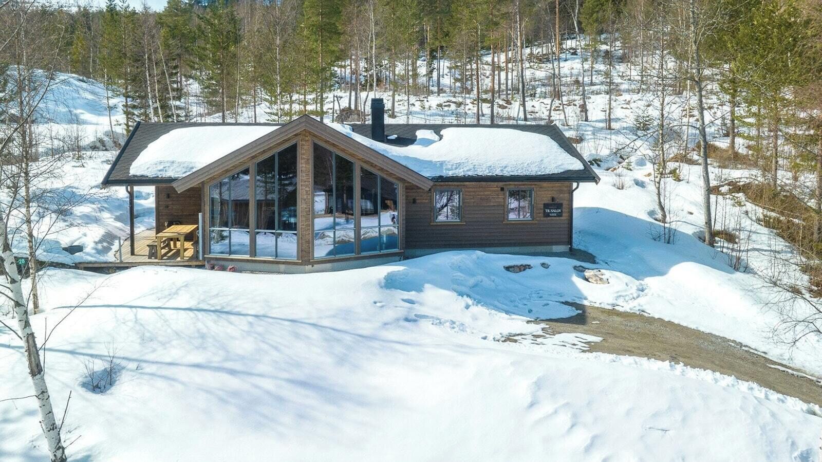 En stuga med en stor glasfasad, en träpanelbeklädd utsida och ett snötäckt tak.