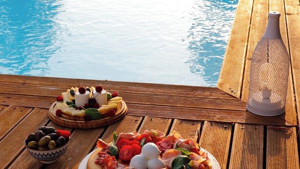 Um deck de madeira com piscina, duas pizzas, azeitonas e uma garrafa de água.
