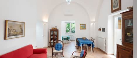 Un vaste espace de vie comprenant un canapé rouge, une table basse en verre, des fauteuils en osier, une table à manger recouverte d’une nappe bleue et un lustre.