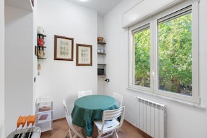 A dining area with a table and chairs, a window with a view of greenery, and shelves with kitchenware and framed botanical illustrations.