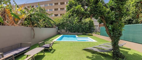 Zona de piscina con césped artificial, sillas de descanso y un edificio al fondo.
