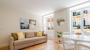 A modern living room with a beige sofa, white chairs, a glass-top table, and a large window with a view of a building. - Cozy Santa Catarina Studio by HostWise (Porto)