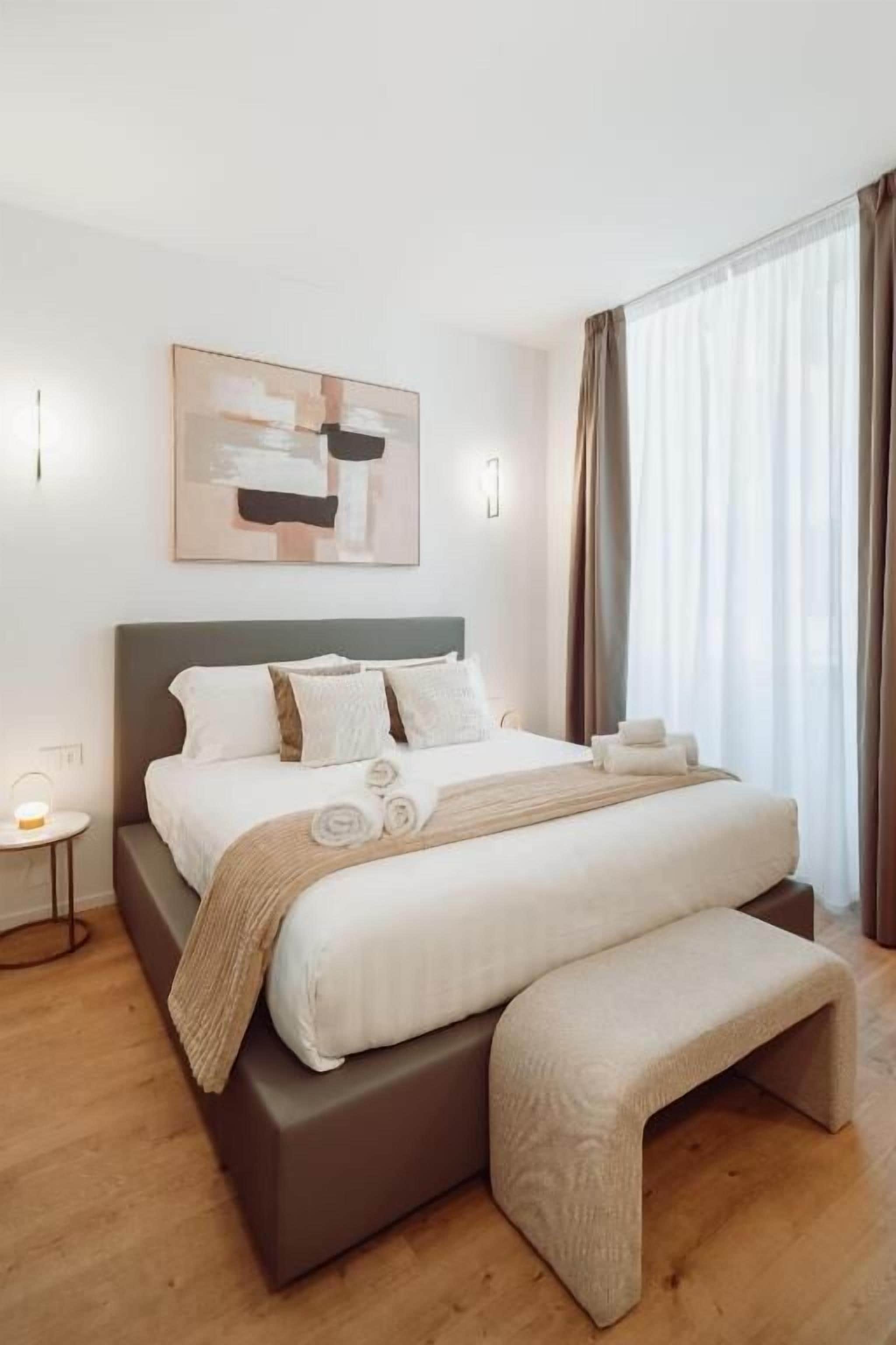 A modern hotel room with a large bed, a bedside table, a bench, and a window with curtains.