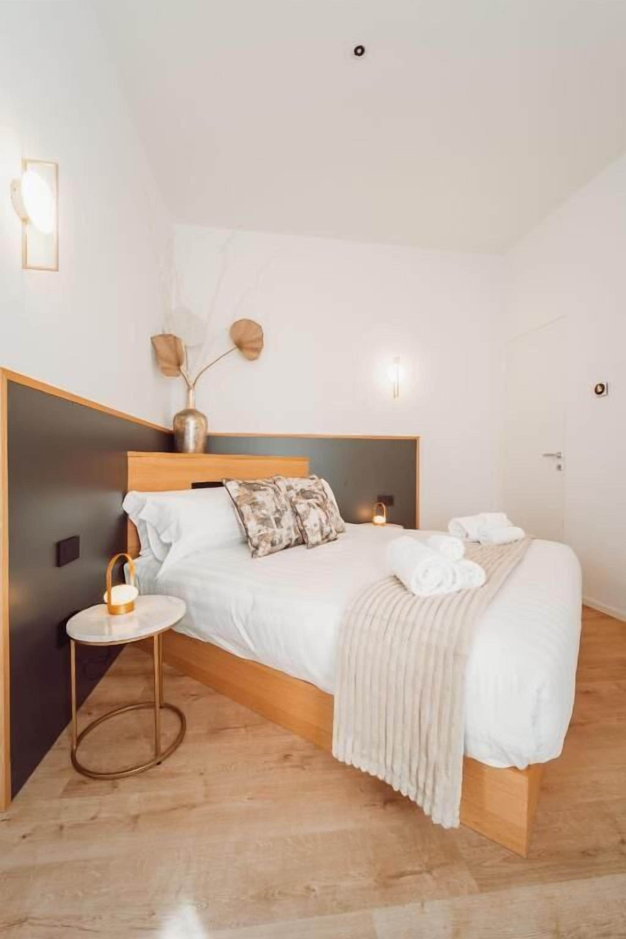 Un dormitorio con cama de madera, ropa de cama blanca, una mesita de noche con lámpara y un jarrón con flores secas en la mesita.