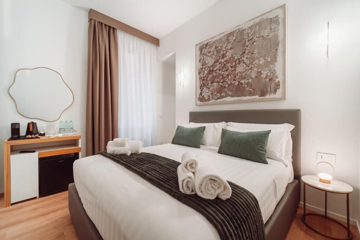 Habitación de hotel con cama, escritorio con silla, mesita de noche con lámpara y un cuadro abstracto grande en la pared.