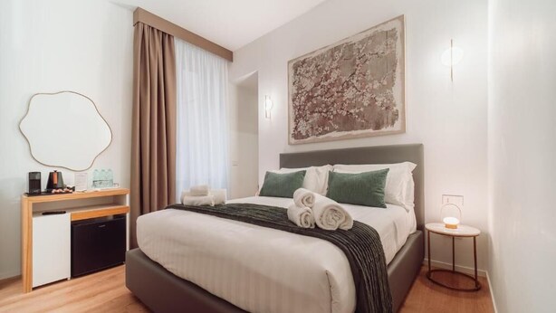 A hotel room with a bed, a desk, a chair, a small table, and a wall-mounted light. - ARCASuite Trevi (Rome)