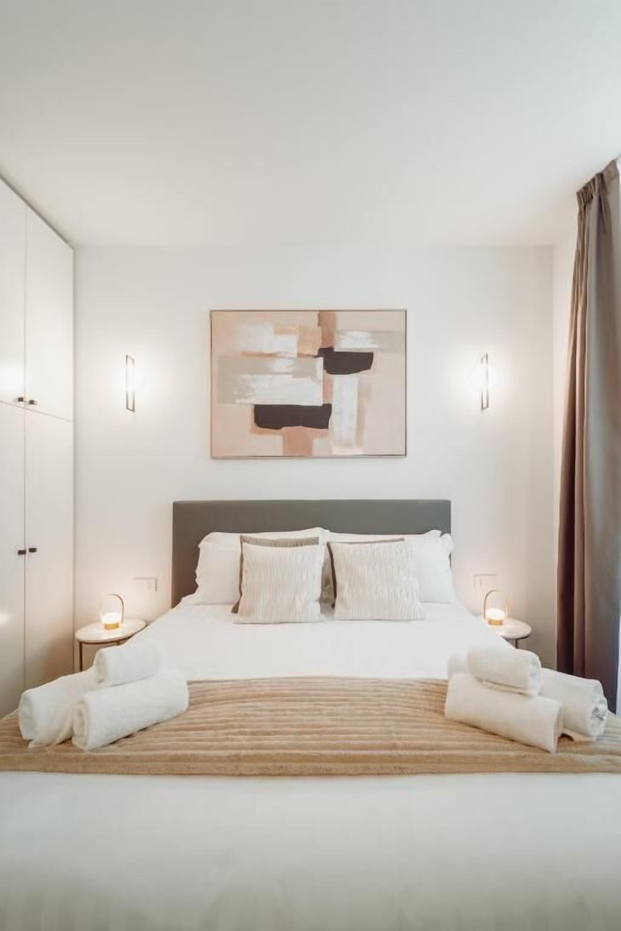 A bedroom with a bed, pillows, a bedside table with a lamp, and a wall-mounted abstract painting.