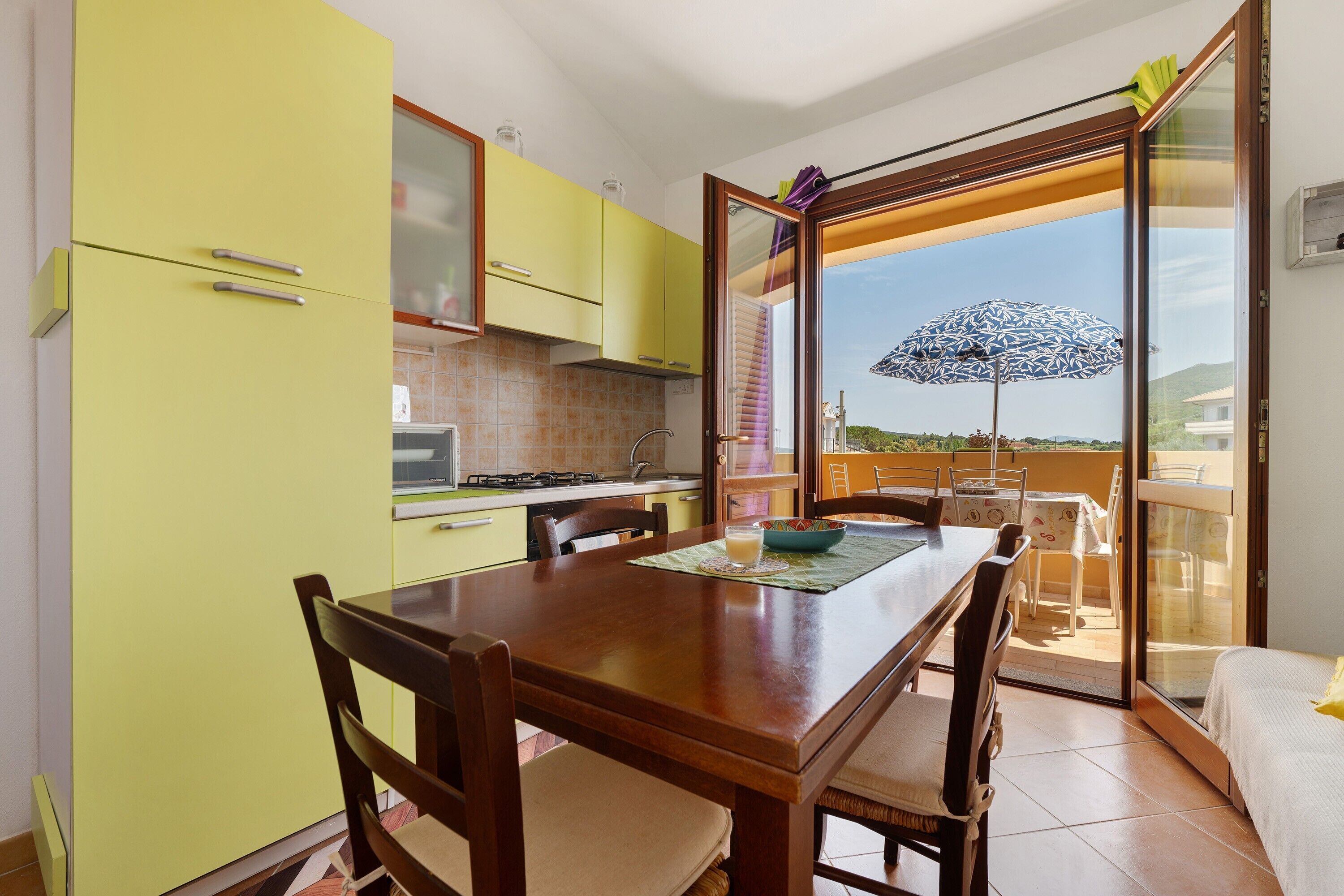A dining area with a wooden table and chairs, a kitchen with yellow cabinets, and a balcony with a patterned umbrella.