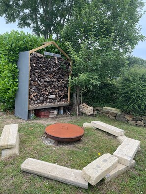 A rustic outdoor setting with a wooden structure containing firewood, a metal fire pit, and stone steps. - "P'ty Penty," a small Breton cottage (Plougastel-Daoulas)